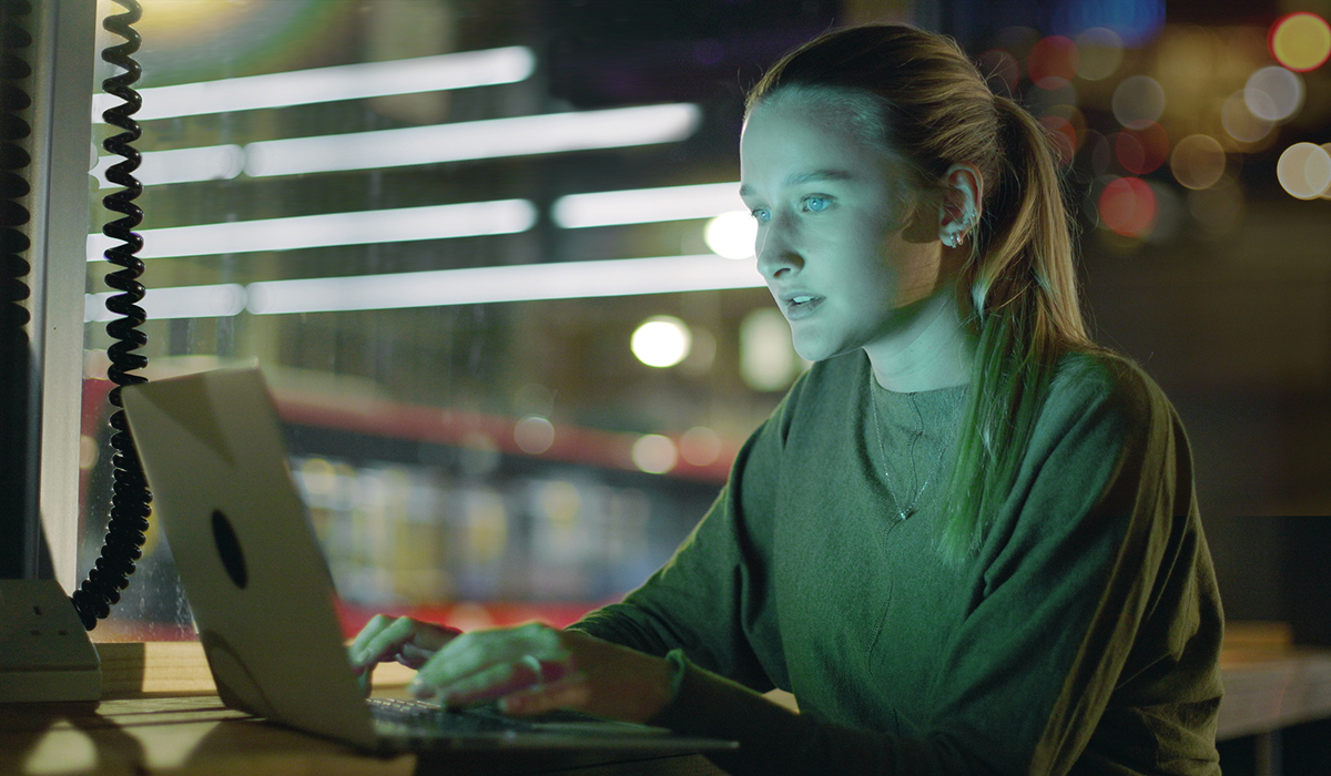 Young woman using a laptop in a hot desk office at night, with city life going on in the background
