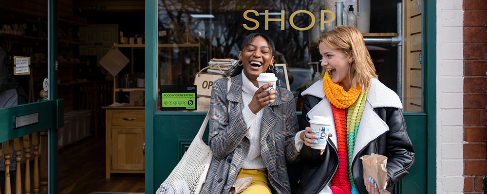 Two friends sitting outside a store, holding coffee and laughing with each other, in the North East of England. Two friends sitting outside a store, holding coffee and laughing with each other, in the North East of England.